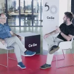 Carlos Iglesias y Pere Tura sentados en sillas blancas dentro de un espacio moderno y minimalista de CeGe. Ambos conversan animadamente, gesticulando mientras intercambian ideas sobre tecnología e innovación. Al fondo, se ve una gran cristalera con una oficina y un cartel conmemorativo del 50 aniversario de CeGe.