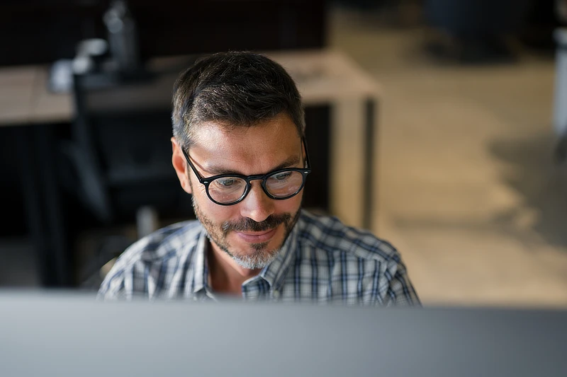 Hombre con gafas y camisa de cuadros mirando atentamente la pantalla de su ordenador en una oficina, analizando datos para la implementación del XBRL en informes de sostenibilidad.