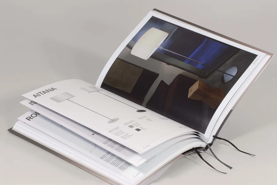 Catálogo impreso abierto con fotografías de lámparas y fichas técnicas, encuadernado en tapa dura, sobre fondo neutro y con varias páginas desplegadas.