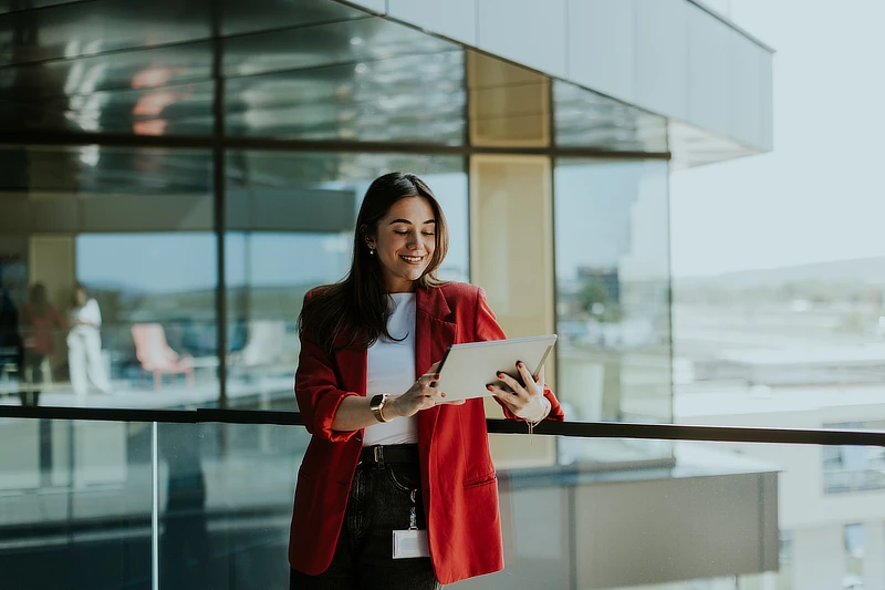 Mujer joven con chaqueta roja utilizando una tableta digital en un espacio de oficinas acristalado.