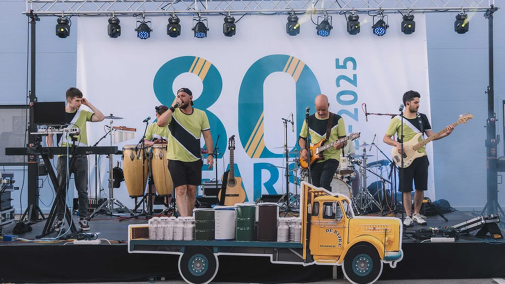 Groupe musical en concert sur scène avec la banderole des 80 ans de De Rijke en arrière-plan.