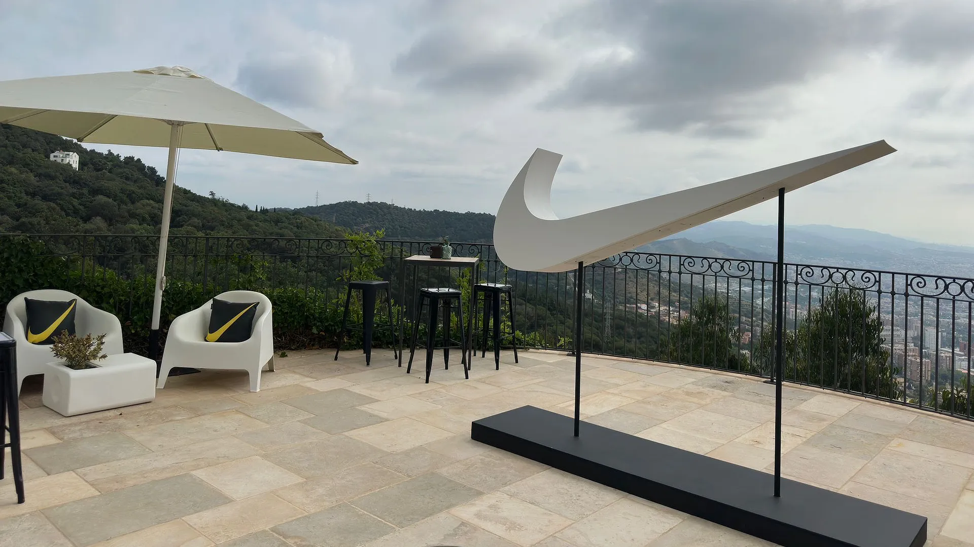 Escultura gran del logotip Nike Swoosh flotant sobre terrassa amb vistes panoràmiques de Barcelona.
