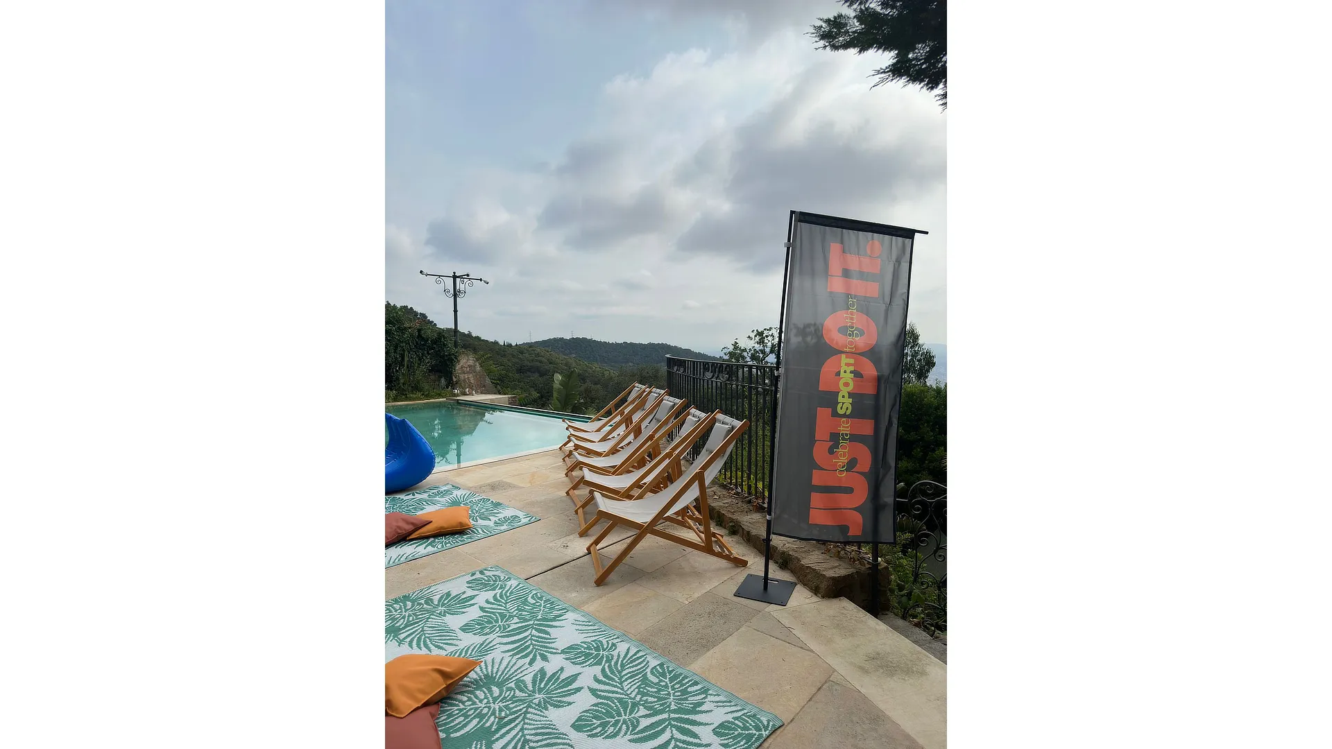 Bandera vertical Nike Just Do It Day col·locada al costat d’una piscina amb zona de descans exterior.