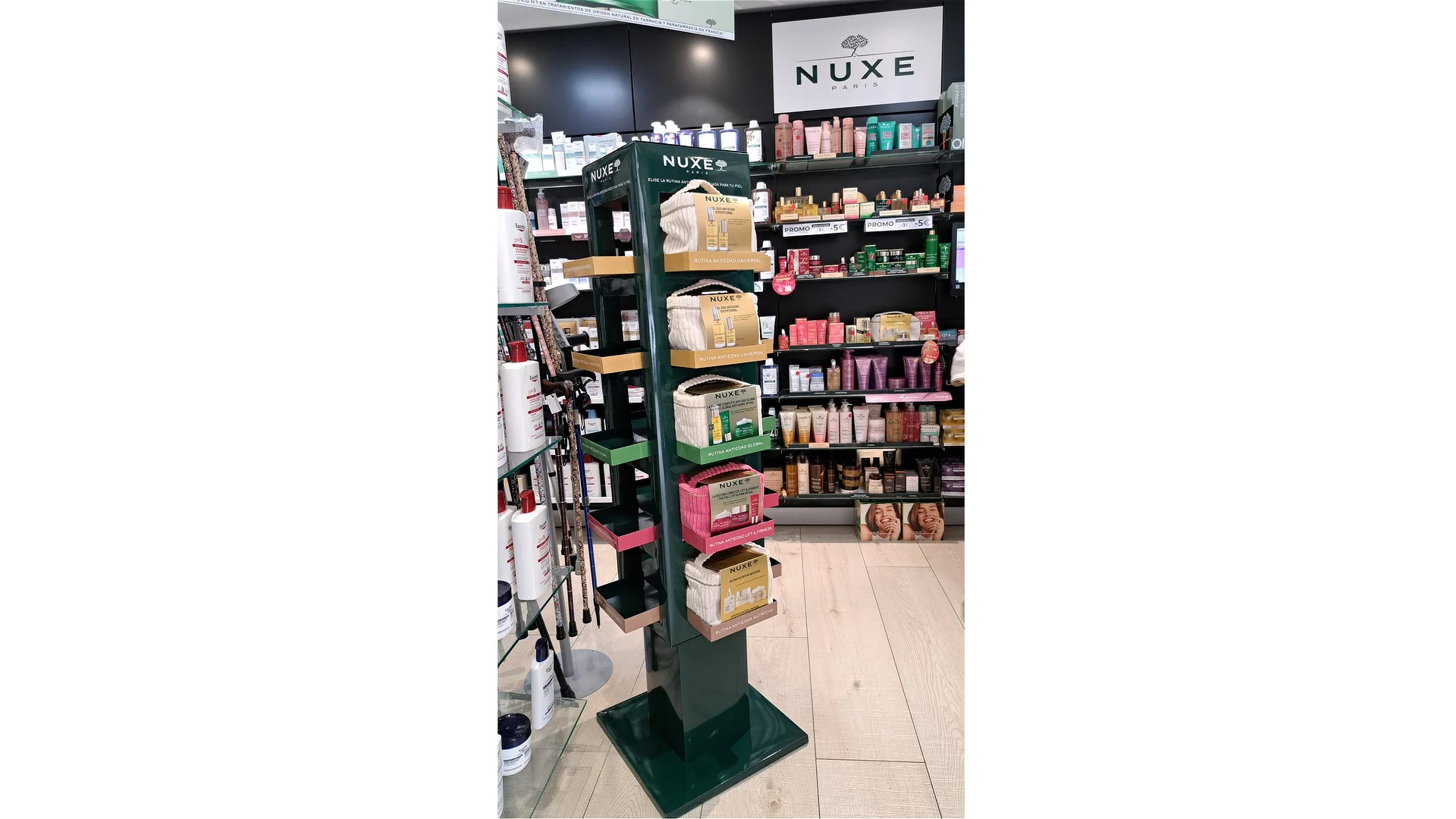 Expositor de PLV para Nuxe instalado en farmacia, con neceseres de varios colores colocados en bandejas verdes y doradas, rodeado de productos cosméticos en estanterías.