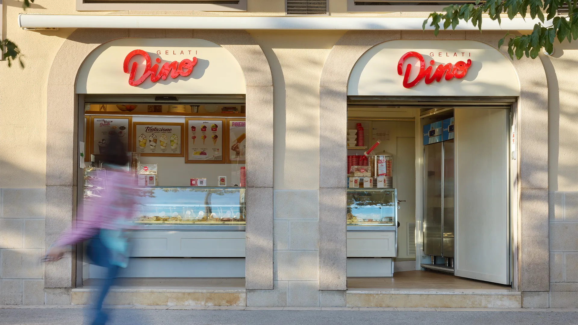 Fachada de tienda Dino Helados con rótulo retroiluminado rojo y vitrinas de helados visibles desde la calle, en un entorno urbano iluminado por la luz del día.