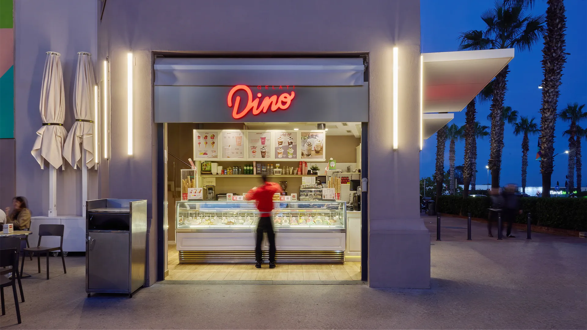 Establecimiento Dino Helados de noche con rótulo rojo retroiluminado, interior visible y ambiente exterior con iluminación cálida y palmeras en el paseo.
