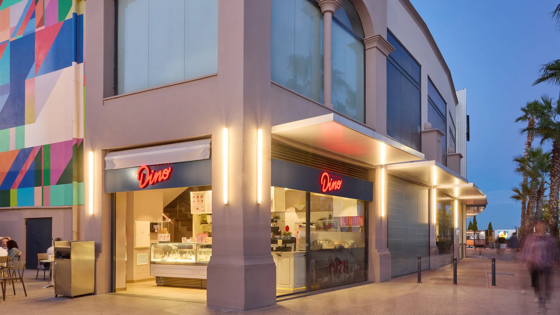 Tienda Dino Helados iluminada al atardecer con rótulos retroiluminados y espacio exterior con terrazas y decoración mural de colores.