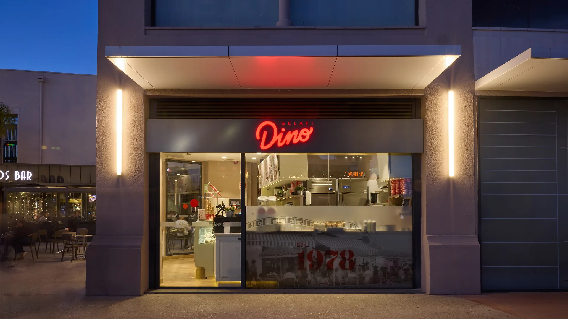Fachada frontal de Dino Helados por la noche con rótulo rojo brillante y cristalera que muestra el interior del local y su actividad.