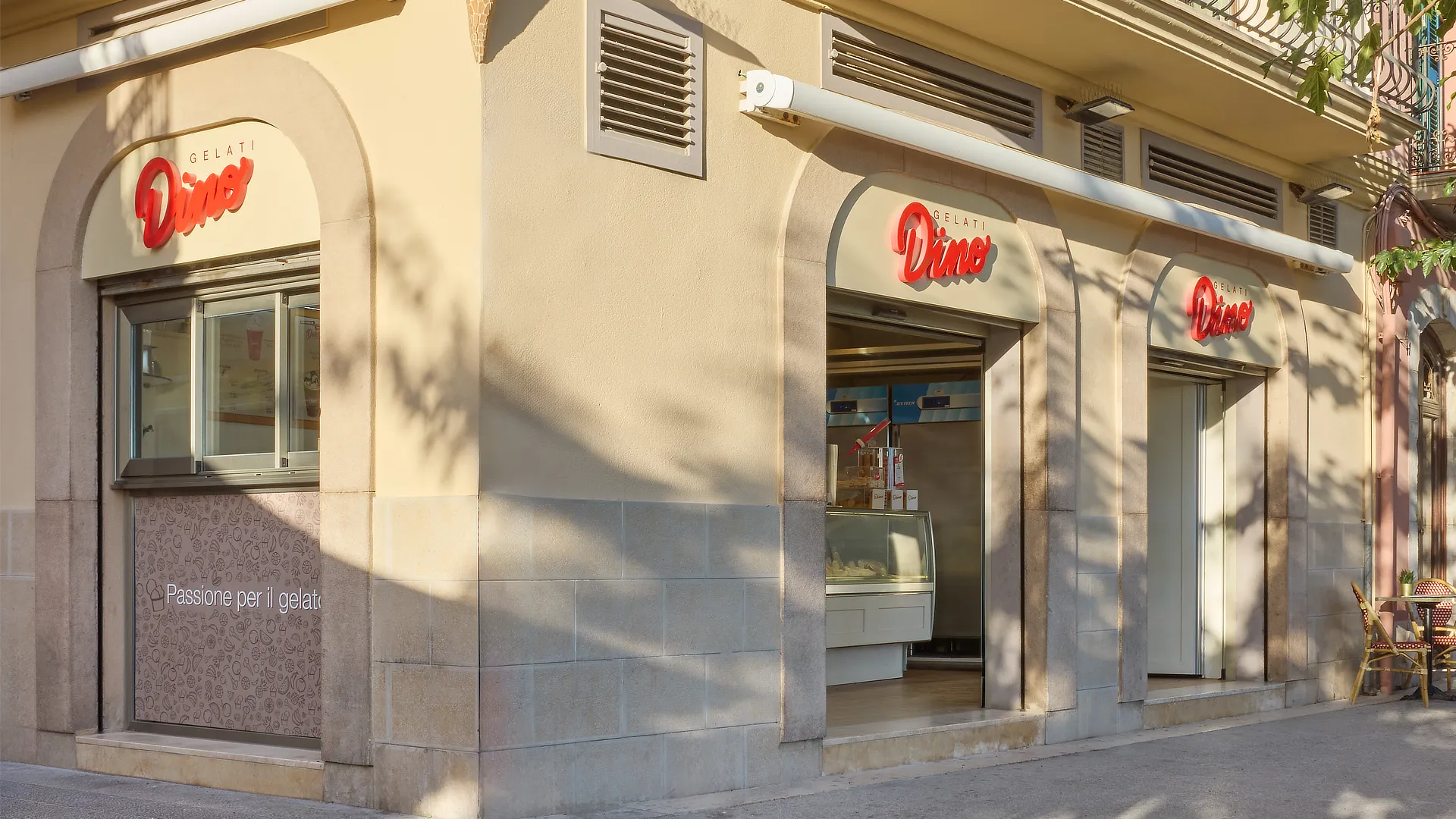 Esquina de tienda Dino Helados con rótulos retroiluminados, persianas metálicas y gráfica inferior “Passione per il gelato” en tonos suaves.