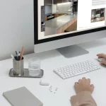 Mesa de trabajo minimalista con ordenador mostrando maquetación de catálogo, teclado, vaso de agua y material de oficina, representando procesos digitales de diseño editorial.