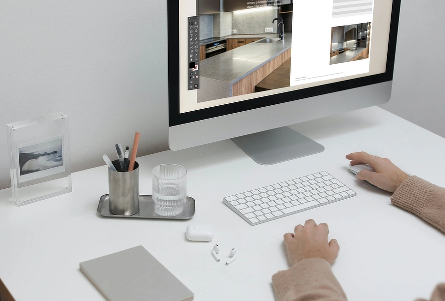 Mesa de trabajo minimalista con ordenador mostrando maquetación de catálogo, teclado, vaso de agua y material de oficina, representando procesos digitales de diseño editorial.