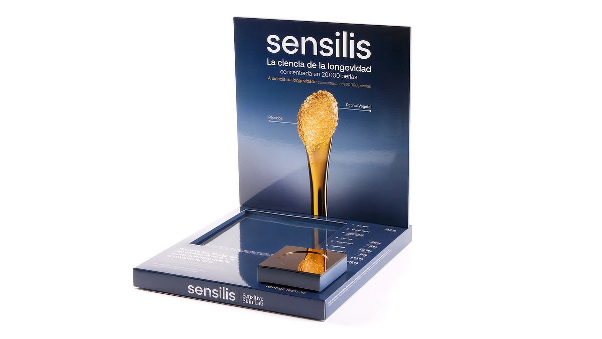 Expositor de Sensilis para farmacias con fondo azul, imagen ampliada de perlas doradas y espacio frontal destinado al producto, diseñado para presentar la nueva crema antiedad.