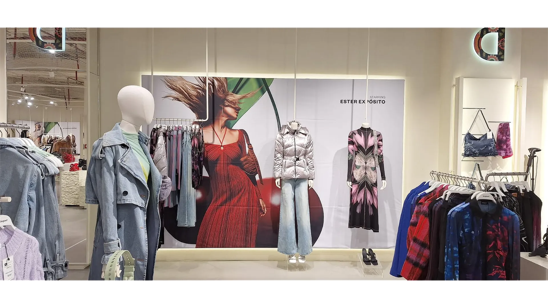 Interior de tienda con panel retroiluminado de la campaña gran formato Desigual mostrando a una modelo, rodeado de maniquíes, prendas expuestas y una composición visual que resalta la creatividad de marca.