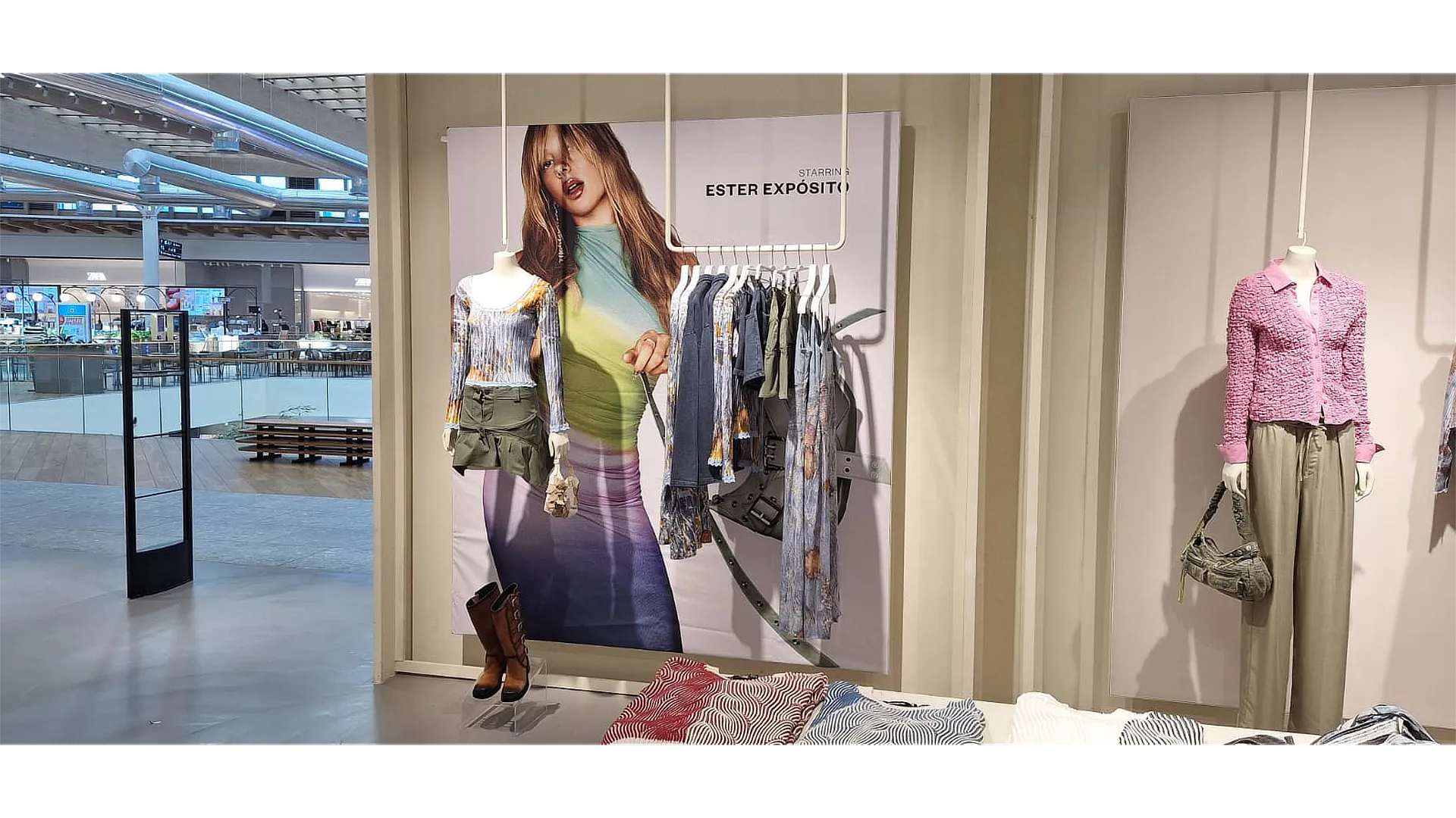 Interior de tienda Desigual con gran panel vertical de campaña gran formato Desigual mostrando a Ester Expósito, acompañado de prendas expuestas y mobiliario comercial iluminado.
