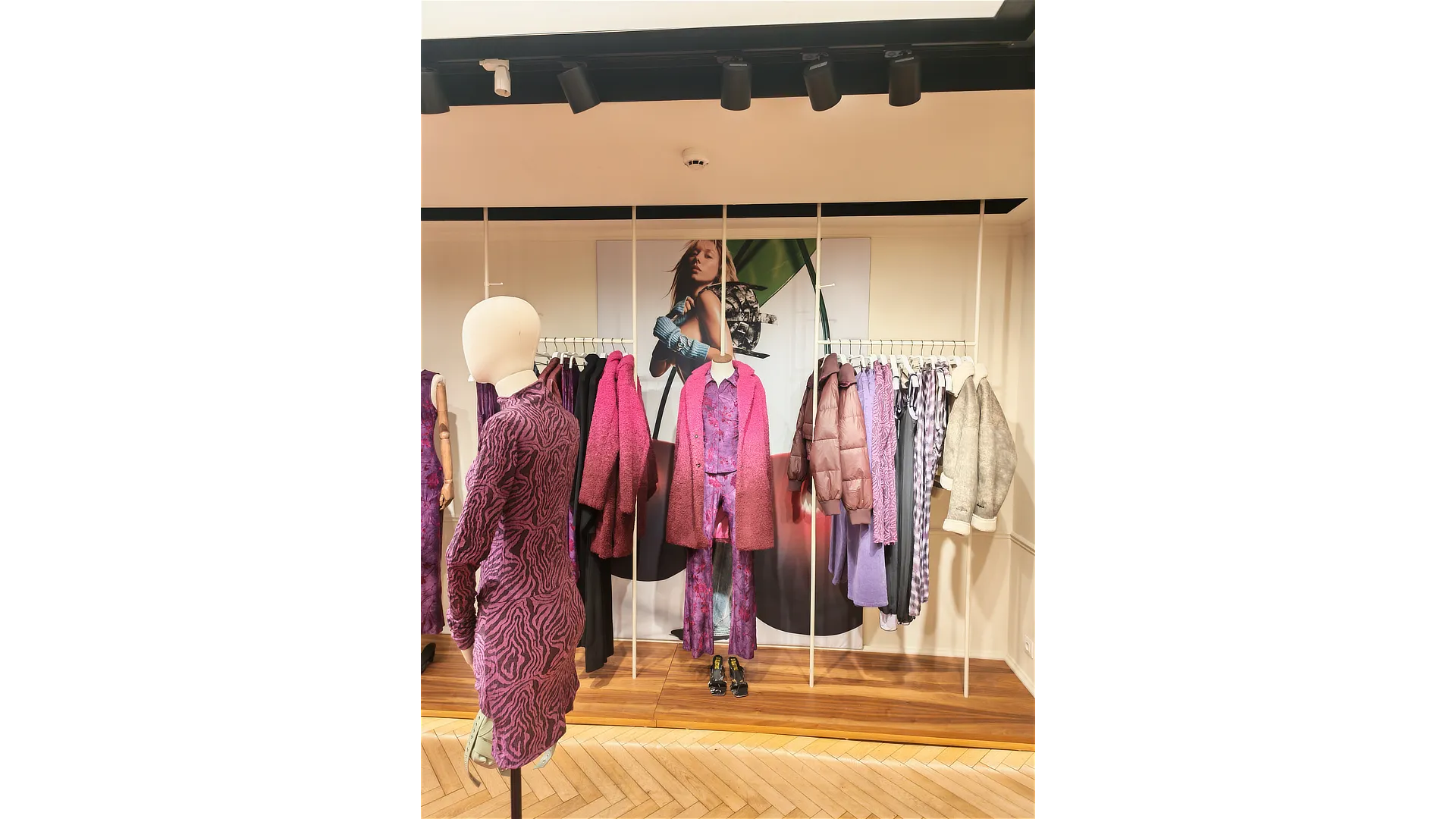 Interior de tienda con la campaña gran formato Desigual, mostrando panel gráfico de Ester Expósito, percheros con prendas en tonos morados y un maniquí en primer plano.