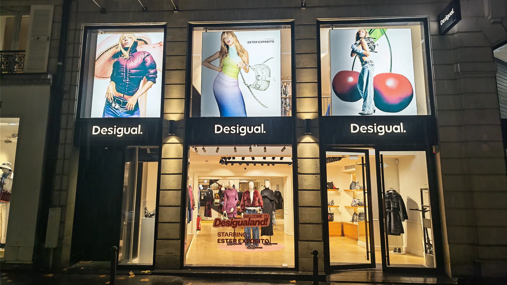 Vista frontal de tienda Desigual con paneles de campaña gran formato Desigual en la parte superior y escaparte con mensaje “Welcome to Desigualand” protagonizado por Ester Expósito.