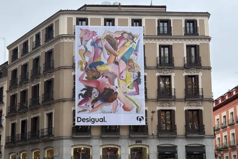 Bâche publicitaire grand format sur la façade d'un bâtiment classique, montrant une illustration artistique colorée de figures féminines et le logo de la marque Desigual.