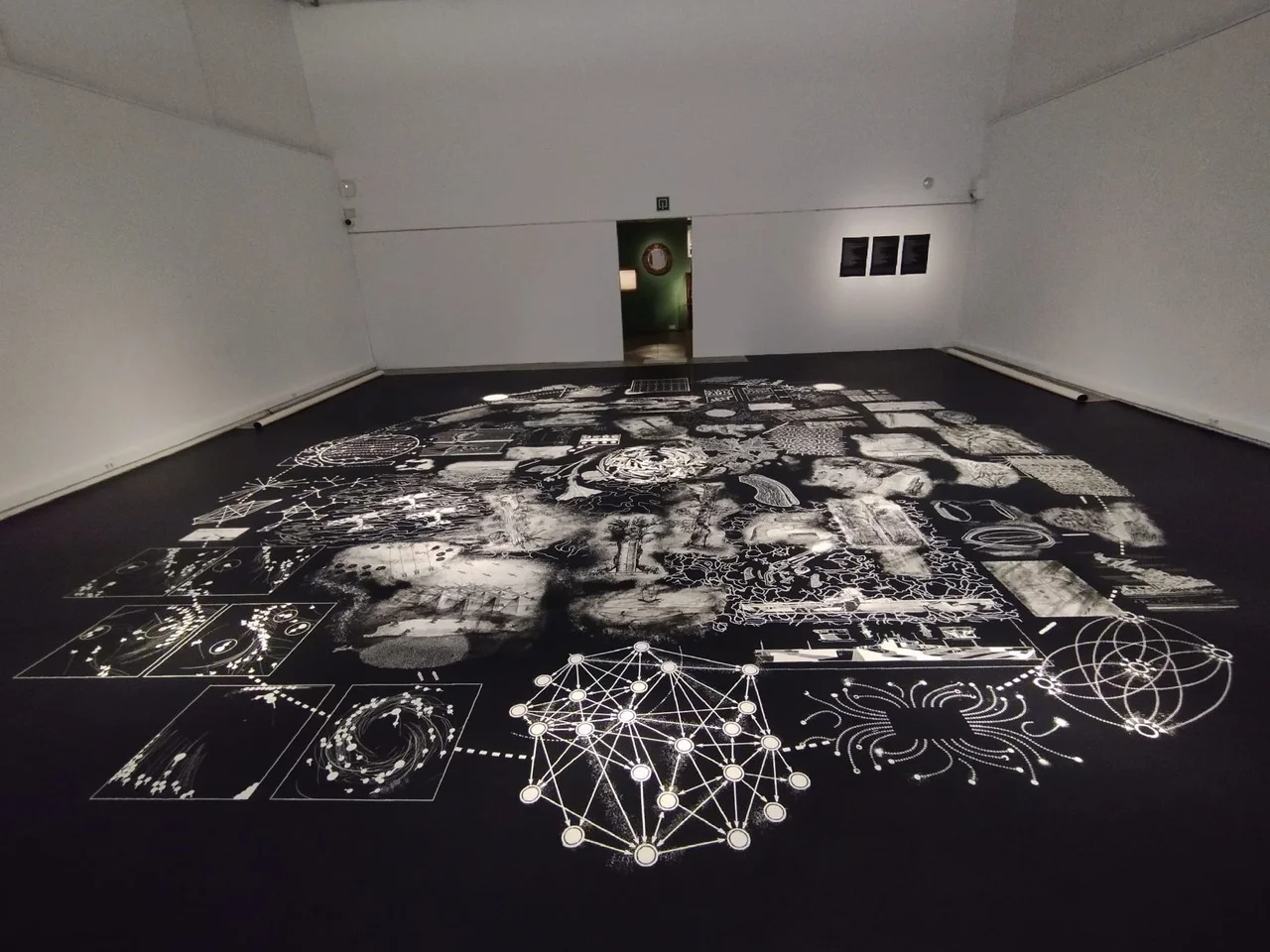 Fotografía frontal de la intervención gráfica en el suelo ocupando gran parte de la sala, con esquemas de constelaciones y redes neuronales impresos.